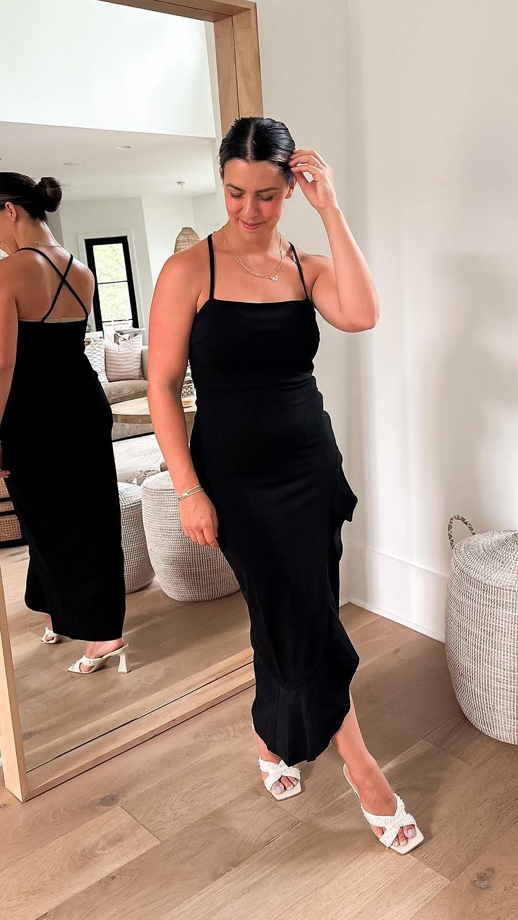 Woman in a black midi dress and white heels posing by a mirror.