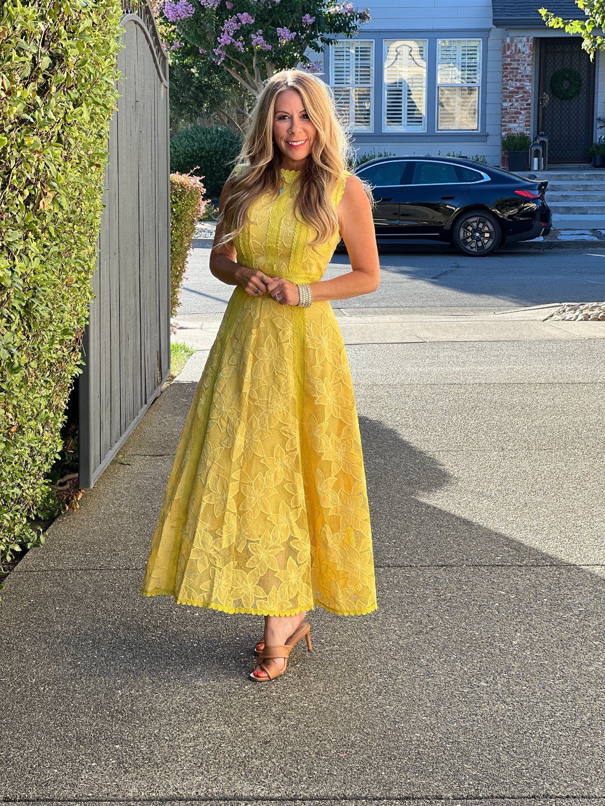 Woman wearing a yellow lace midi dress and tan heels on a sunny driveway.