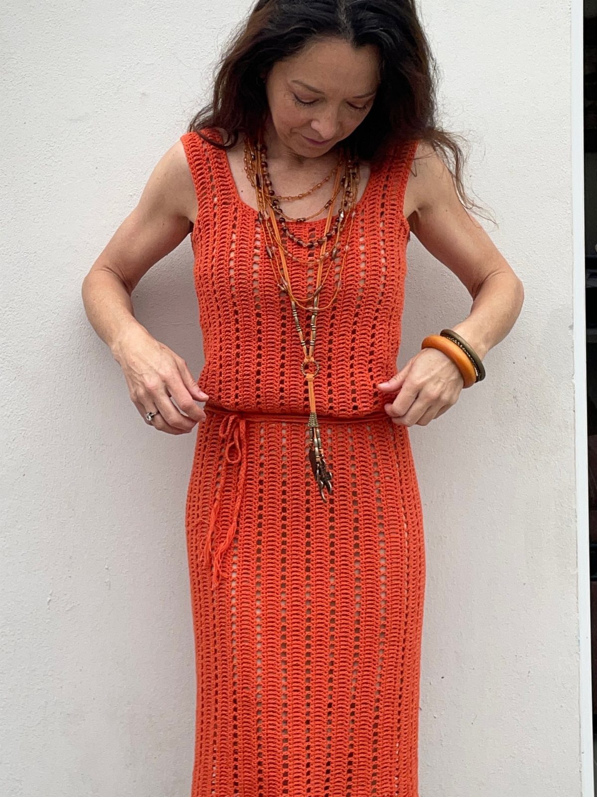 Are Summer Tank Dresses the Season's Must-Have? 8 Styles 7 Woman wearing a long orange crochet tank dress with layered necklaces against a white wall.