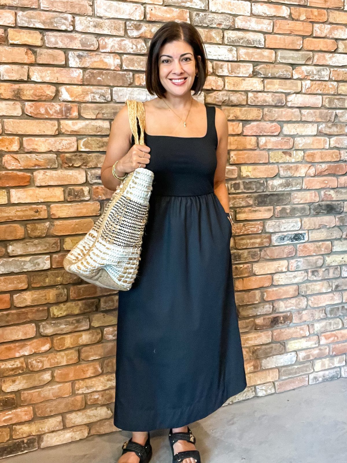 Are Summer Tank Dresses the Season's Must-Have? 8 Styles 1 Woman in a black midi tank dress holding a woven straw bag by a brick wall