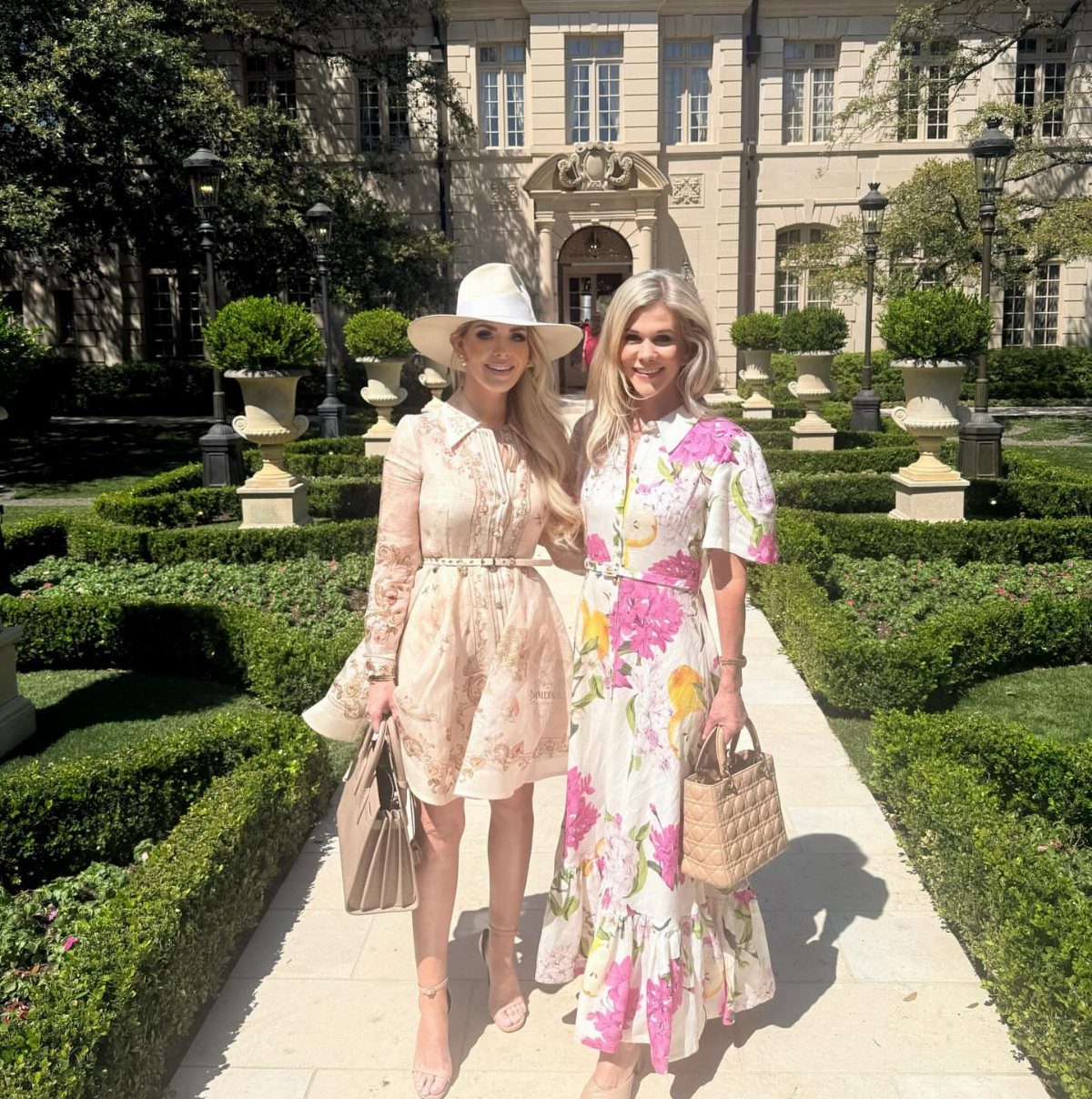 Two women wearing floral dresses and accessories posing in a manicured garden setting.