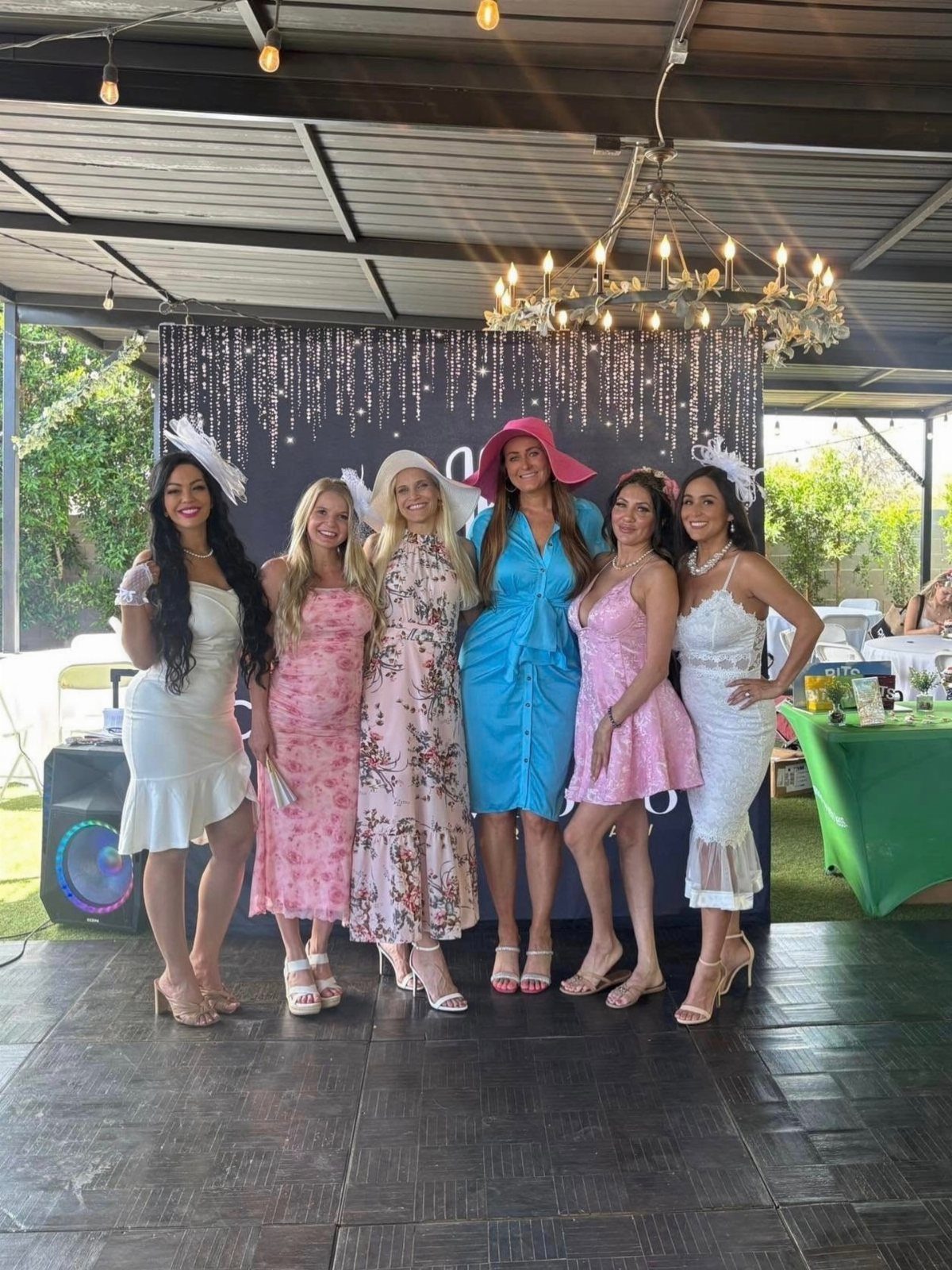 Six women in pastel and white tea party dresses posing before a sparkling backdrop.