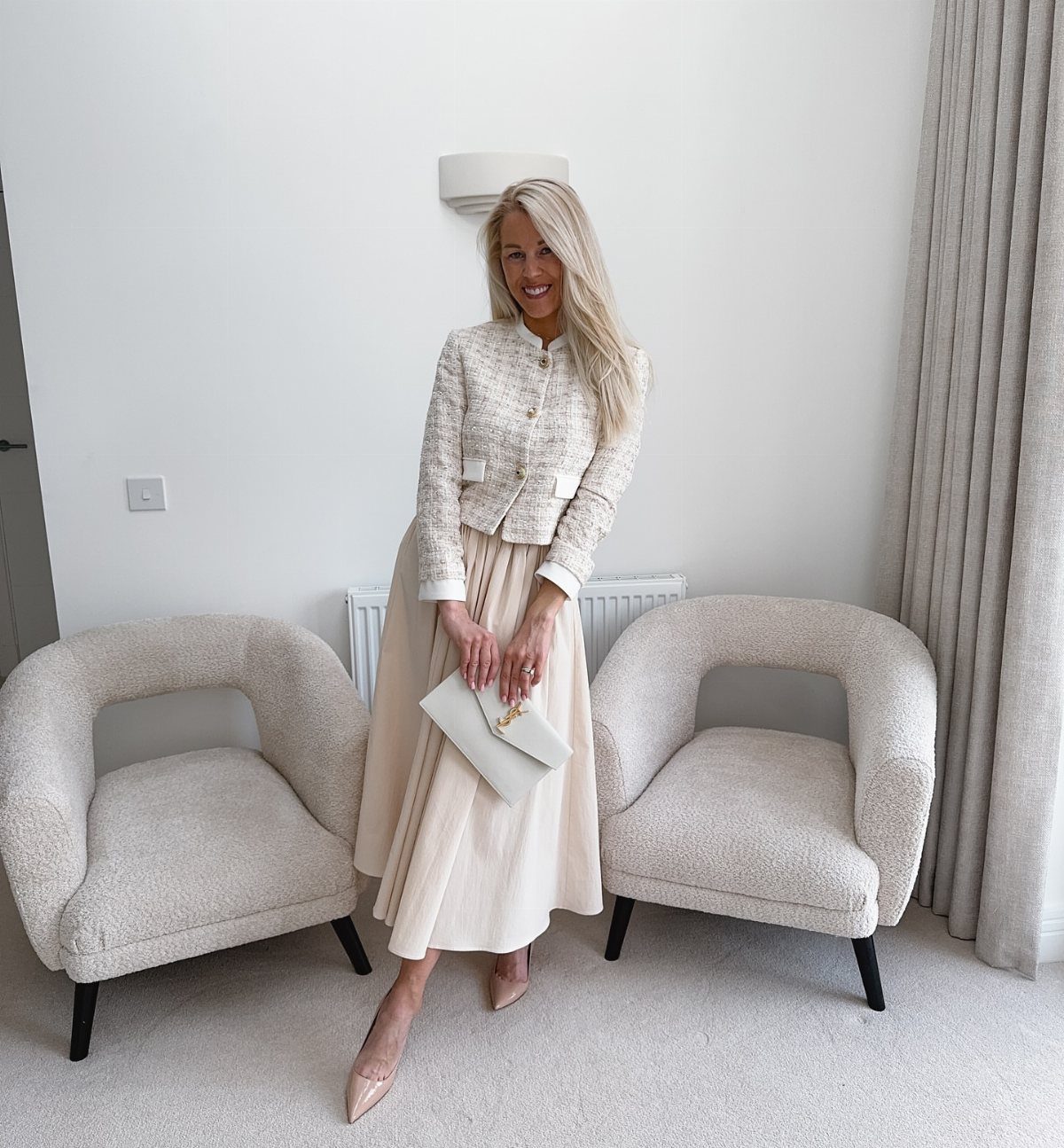 Woman wearing a white tweed structured jacket and cream pleated midi skirt with heels.