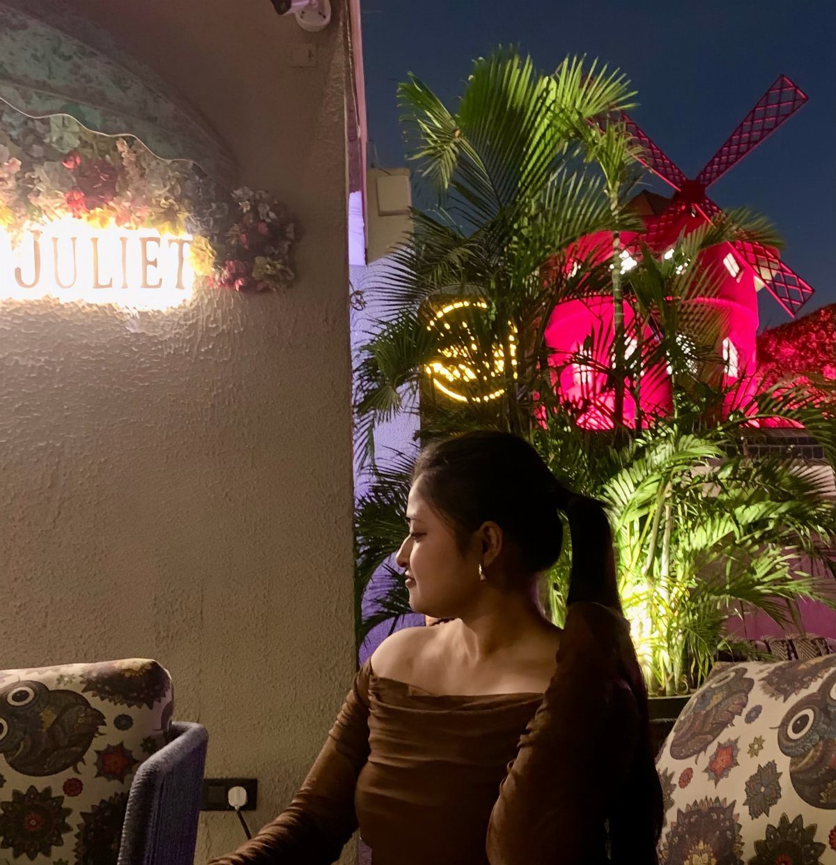 Woman in a brown off-the-shoulder dress sitting outdoors near a glowing sign.