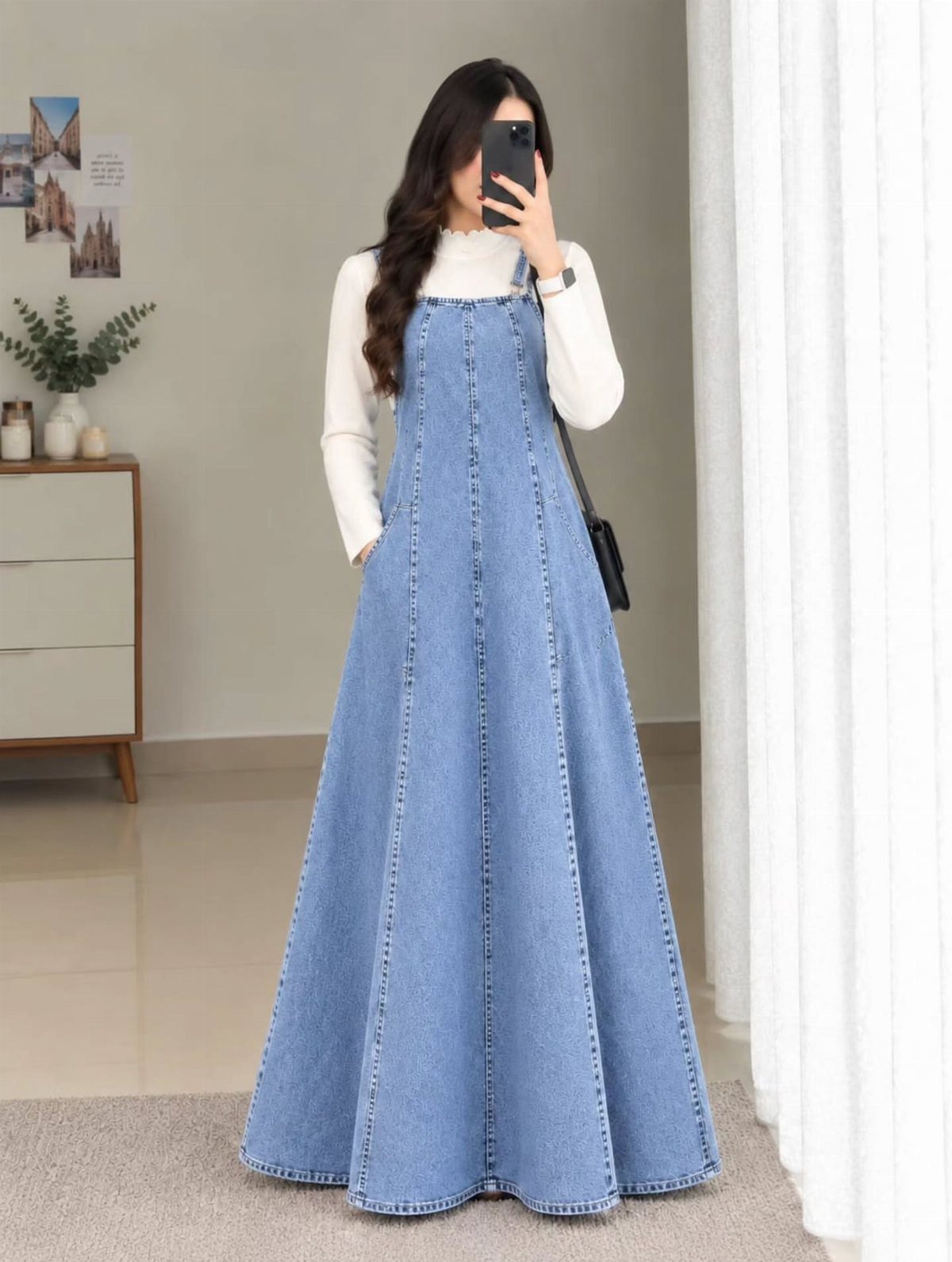 Woman wearing a long blue denim pinafore dress over a white long-sleeved shirt.