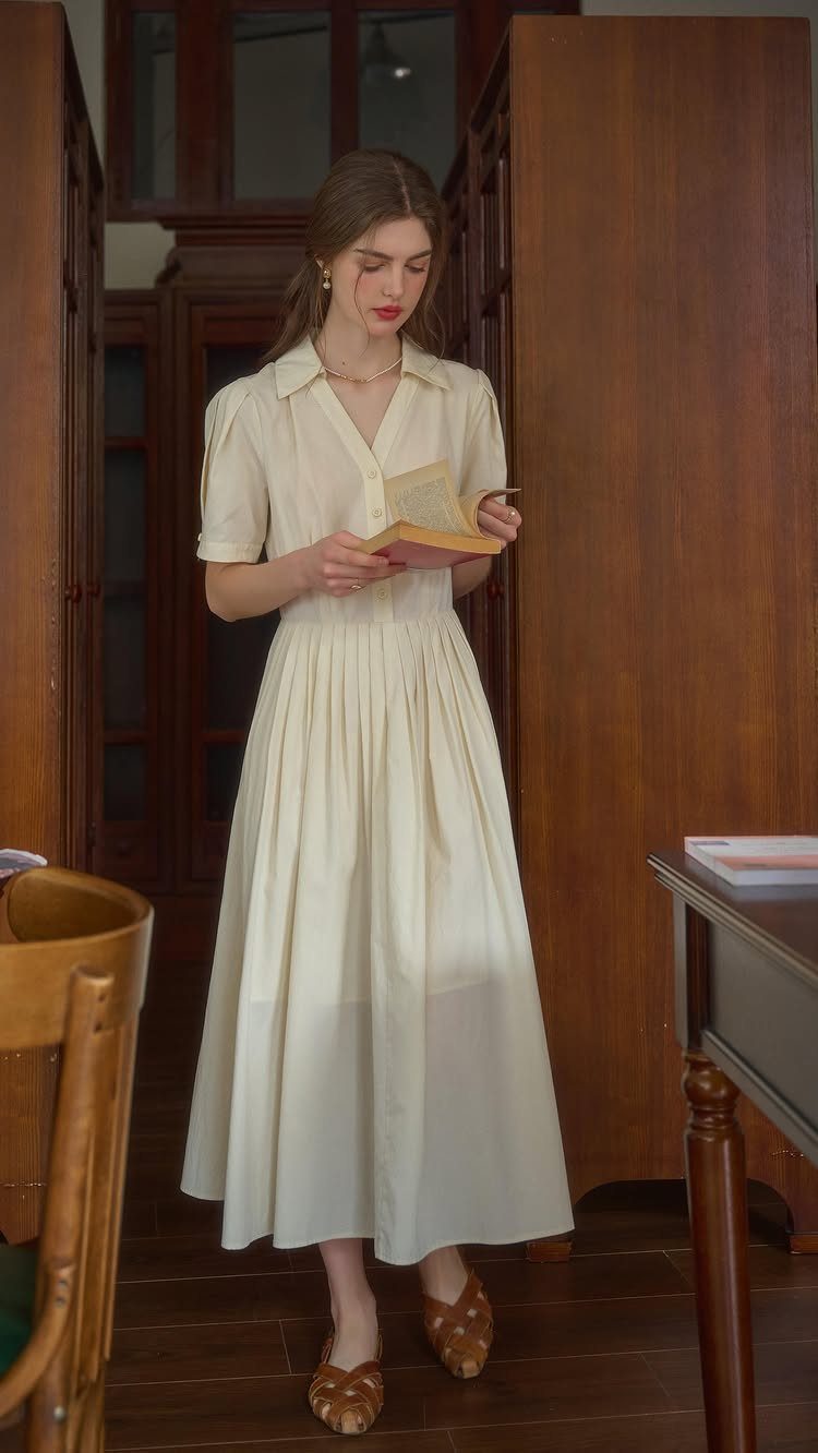 Woman in a cream collared midi dress reading a book in a wood-paneled room.