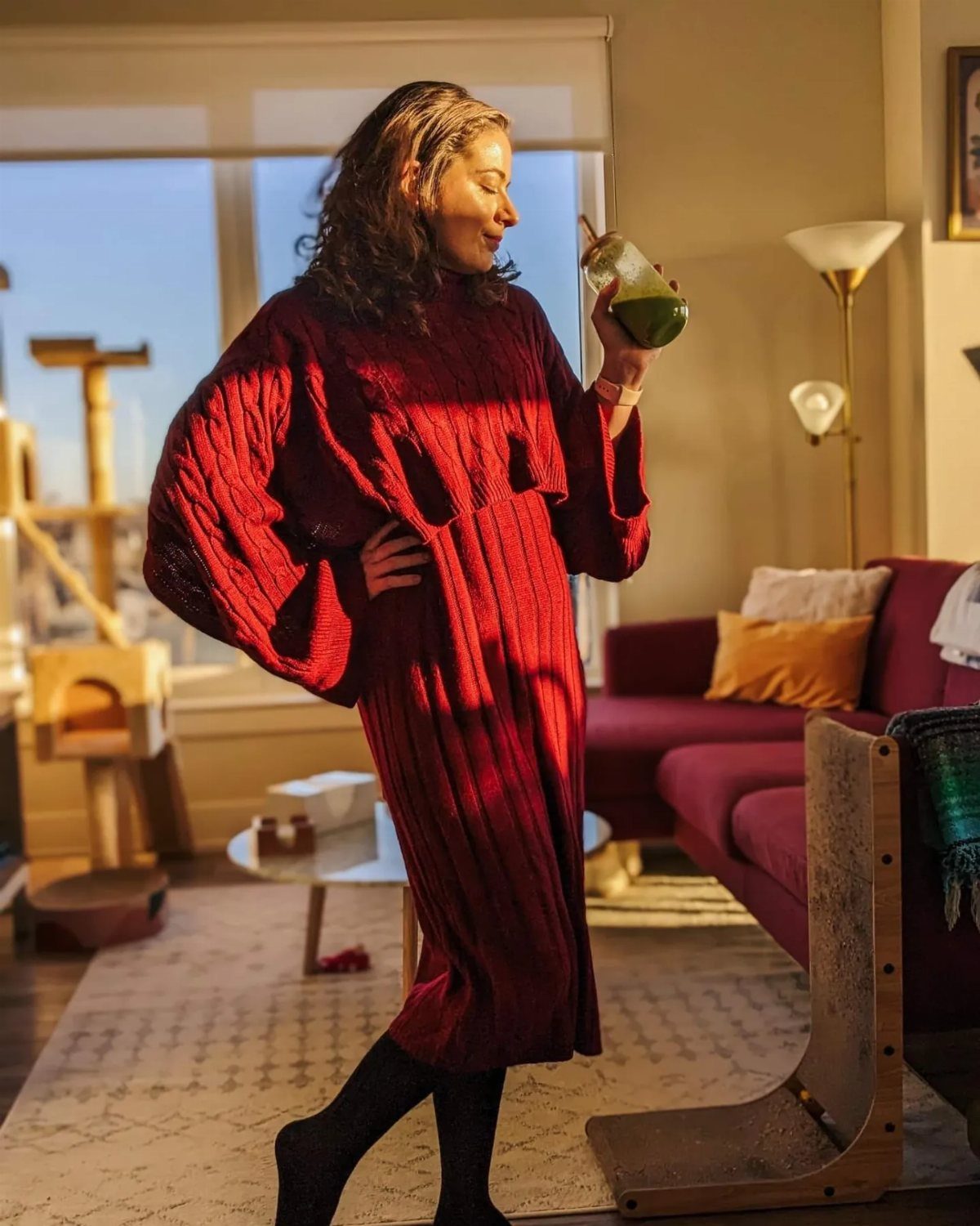 Woman in a chunky red knit sweater dress holding a green drink indoors.