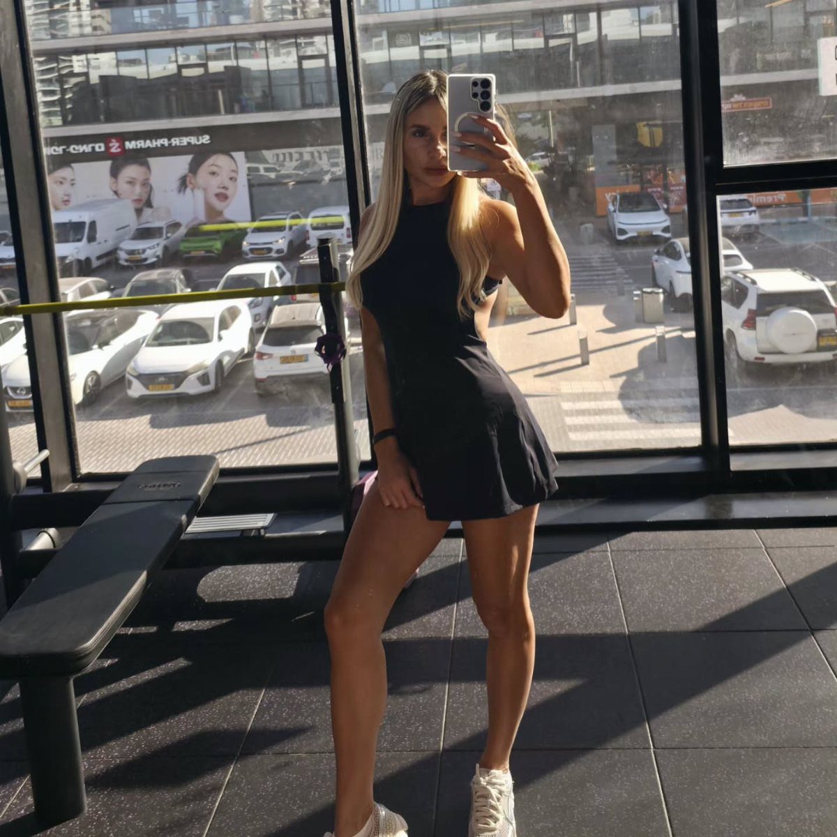 Blonde woman in a black athletic gym dress taking a mirror selfie in a gym.