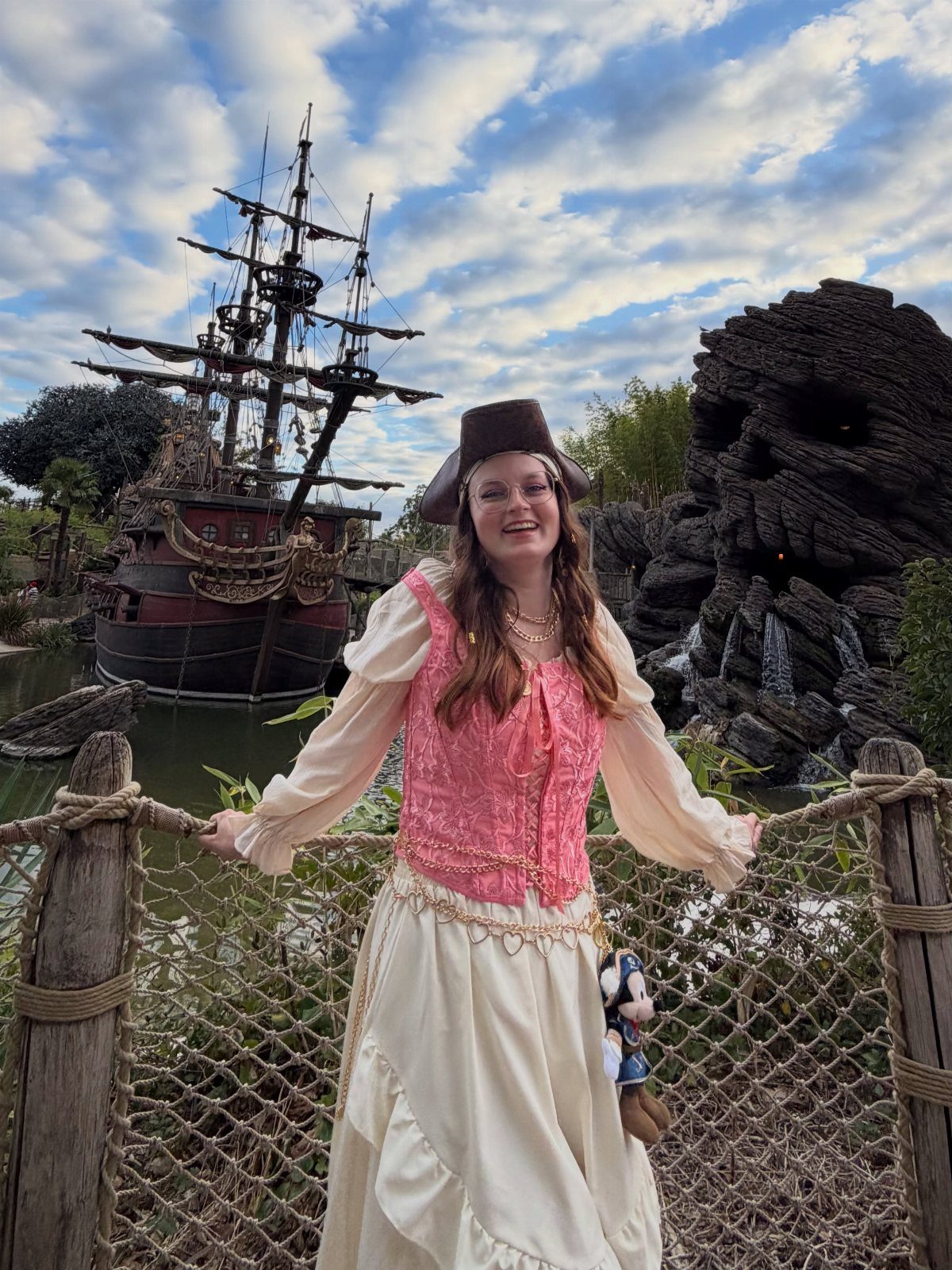 Womens Pirate Dress Styles: 8 Looks from Classic to Bold 8 Woman in a pink and cream pirate dress posing before a pirate ship.